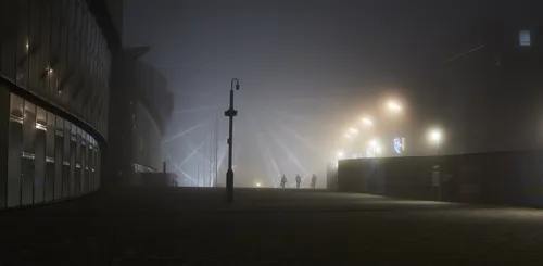 This urban photograph captures the Liverpool wheel, also known as the Liverpool Waterfront Ferris wheel, enveloped in thick fog on an autumn evening in Liverpool, United Kingdom. Illuminated streetlights and building lights are visible through the mist, adding a subdued glow to the scene, while silhouettes of people walk across the paved plaza in front of the wheel. The fog creates a soft veil over the landmarks and modern architecture, contributing to the atmospheric qualities typical of autumn nights in this urban part of Liverpool.